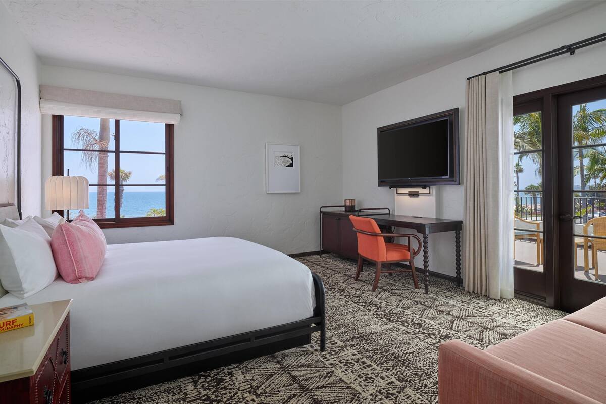 A hotel room with a bed, desk, chair, TV, and a view of the ocean.