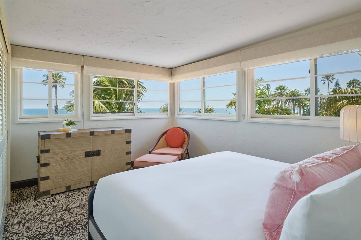 A room with a bed, a chair, a wooden cabinet, and large windows with a view of palm trees and the ocean.