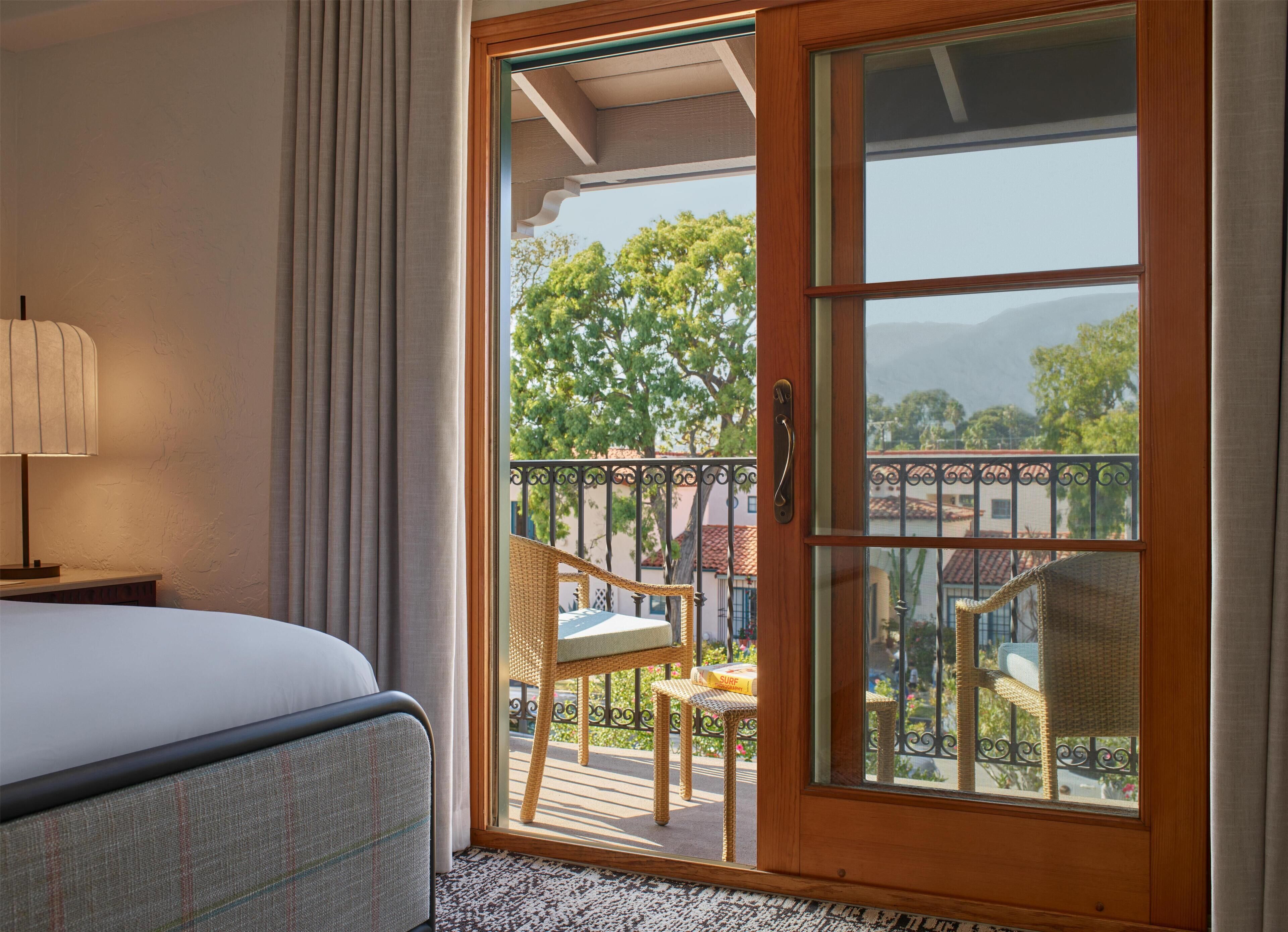 Une chambre d’hôtel avec un lit, une chaise, un balcon aménagé avec des meubles en osier et une vue sur les arbres et les montagnes.