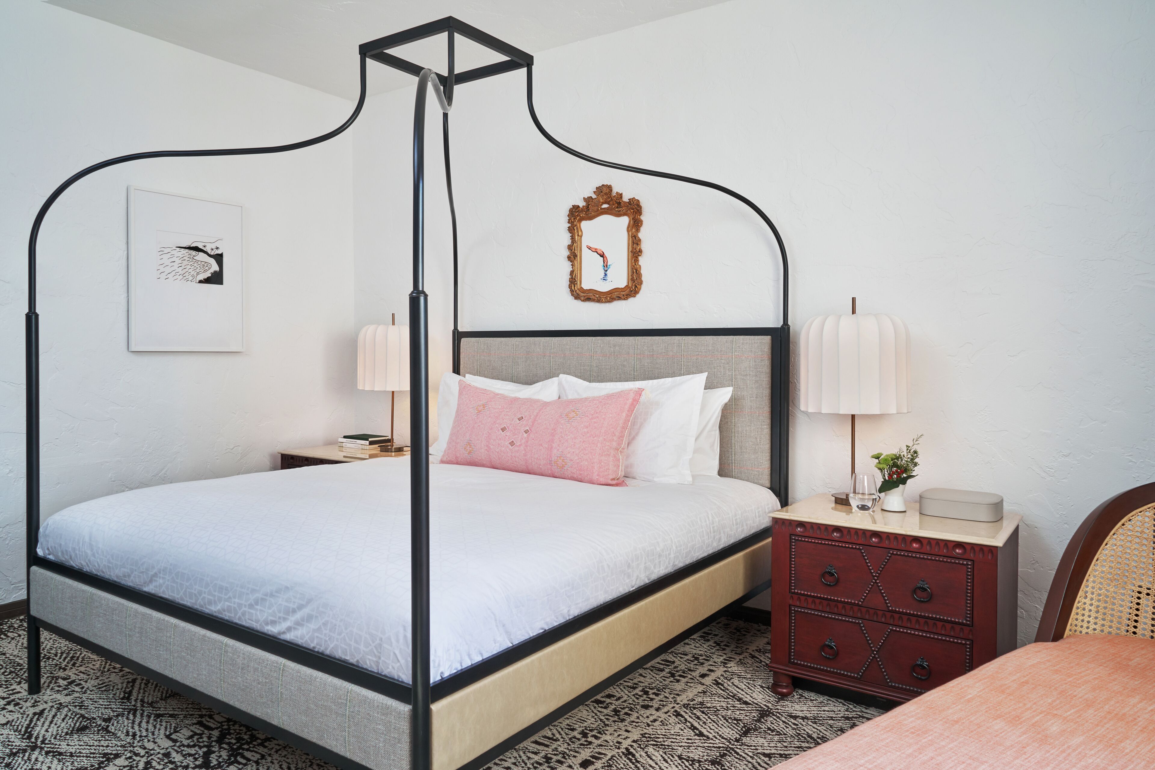 A four-poster bed with a headboard, a bedside table with a lamp, a small dresser, and a framed picture on the wall.