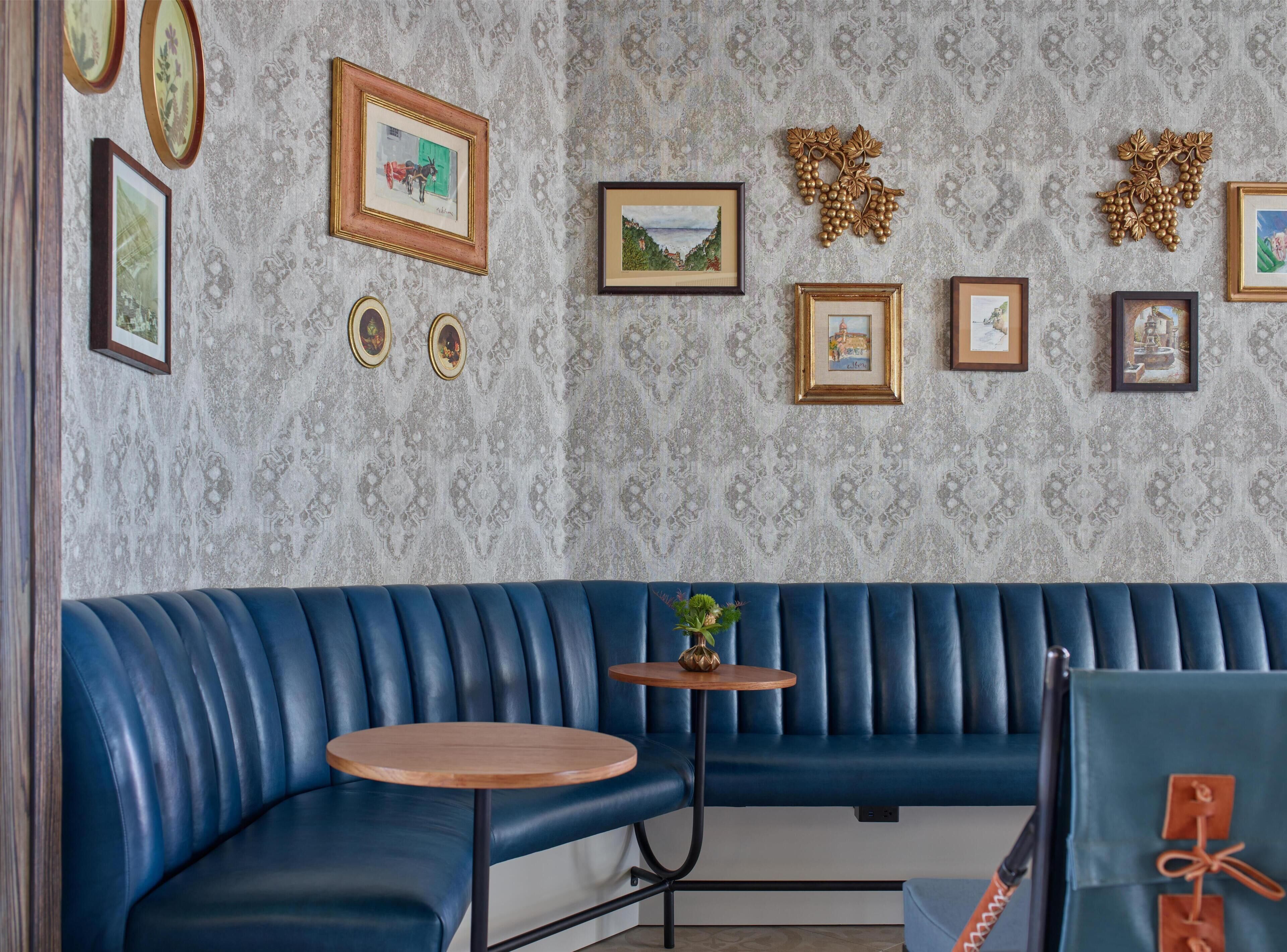 A blue leather booth with a round wooden table, framed pictures on the wall, and a patterned wallpaper.