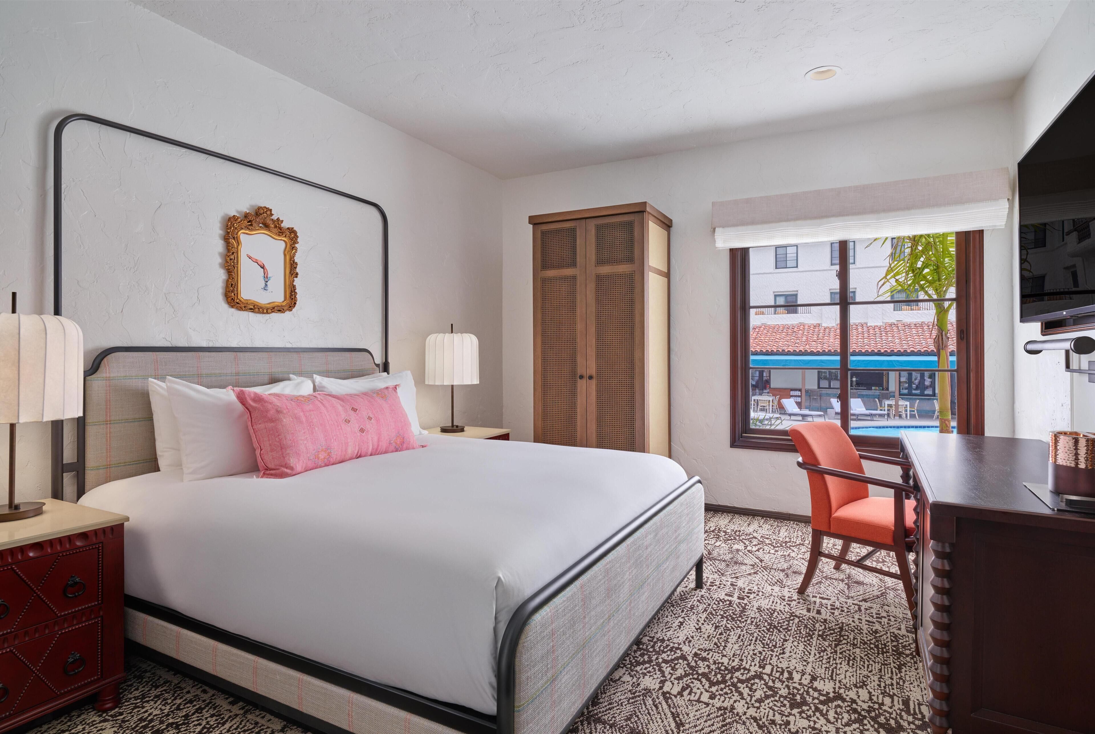 A hotel room with a bed, a desk, a chair, a wardrobe, and a window with a view of buildings and a pool.