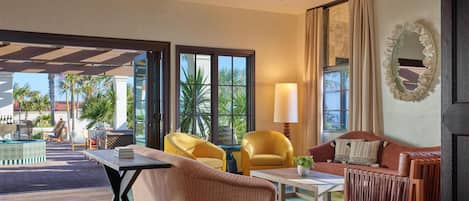 A spacious living area with a sofa, armchairs, a coffee table, and a TV. Large windows offer a view of palm trees and a building outside.