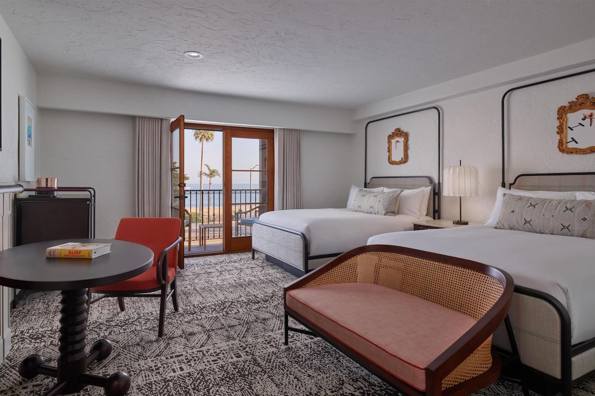 A hotel room with two beds, a desk, a chair, a small table, and a balcony with a view of the beach.