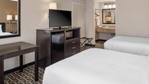 Habitación de hotel con cama, escritorio, televisor, cómoda, espejo y baño con lavamanos y perchero.
