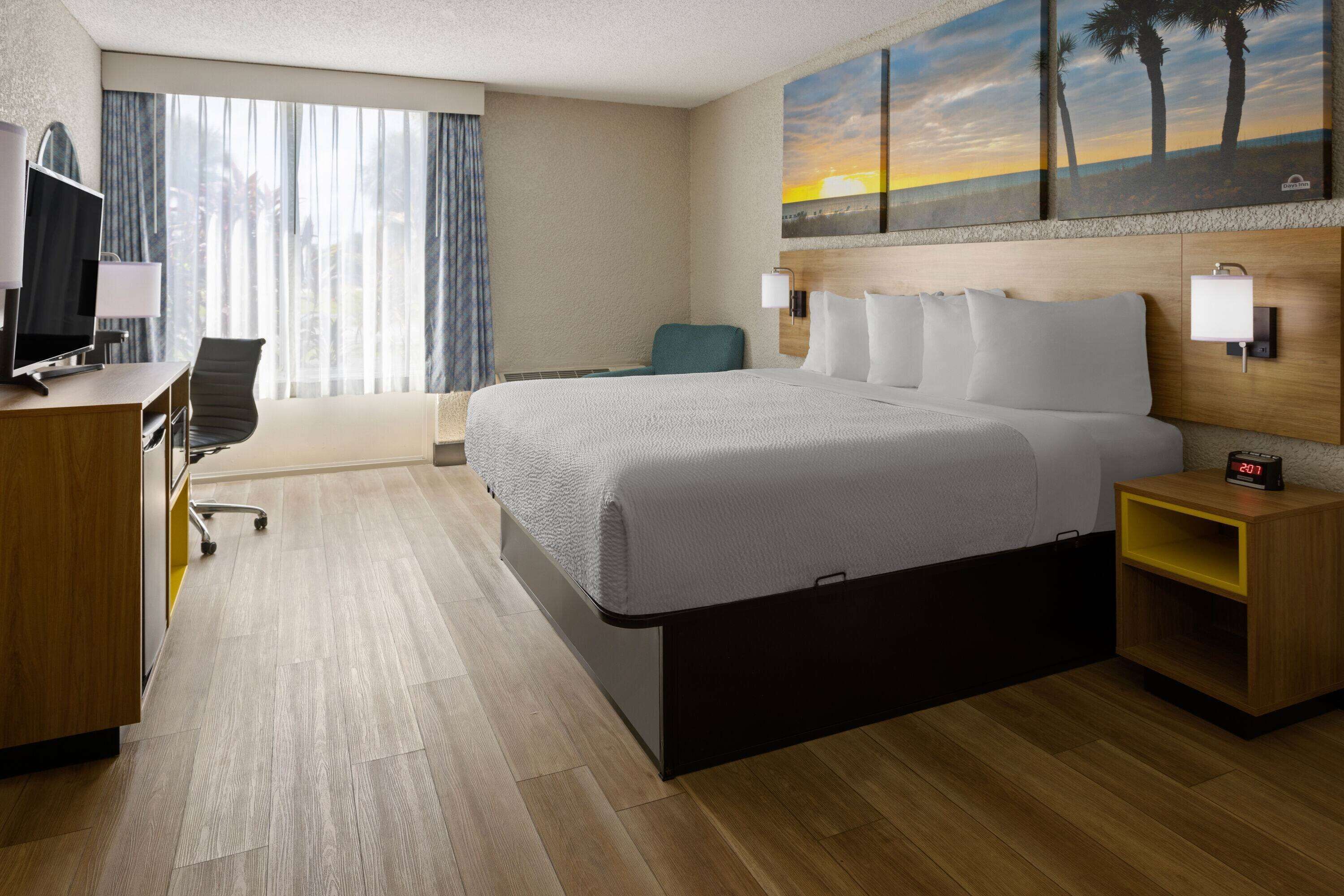Habitación de hotel con cama, escritorio, silla y un cuadro de un atardecer.