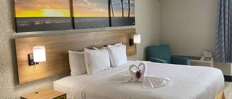 A hotel room with a large bed, a wooden headboard, two bedside tables, a wall-mounted lamp, and a painting of a sunset and palm trees.