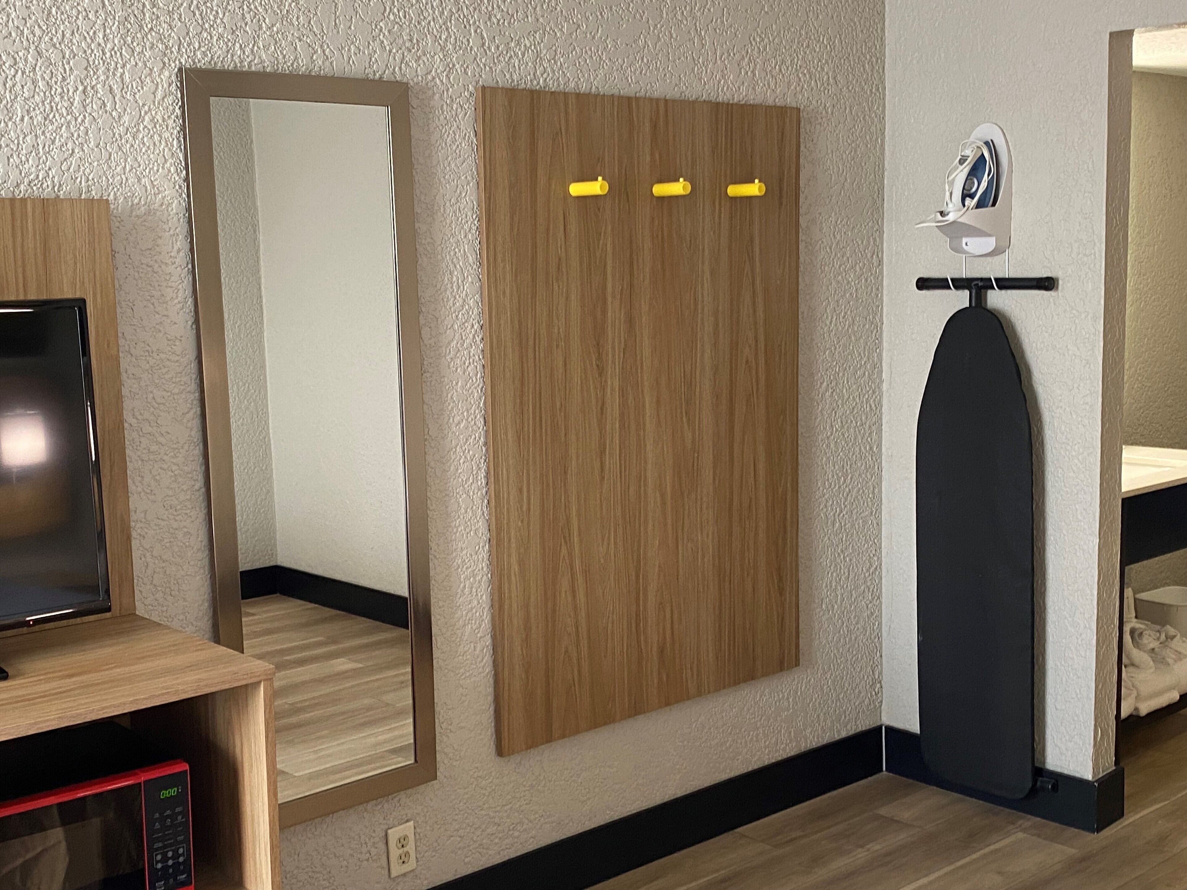 Una habitación con un armario de madera, un espejo, una tabla de planchar negra y un microondas.