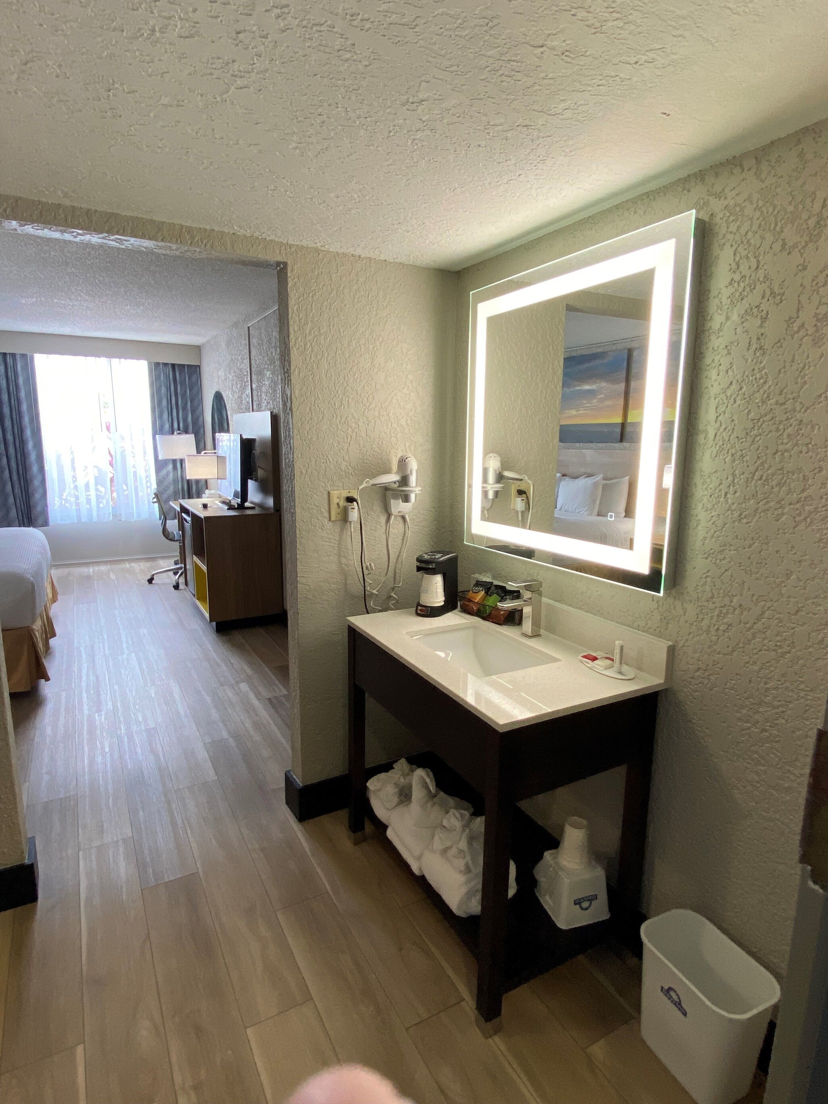 A hotel room with a large mirror, a sink, a coffee maker, and a trash bin.