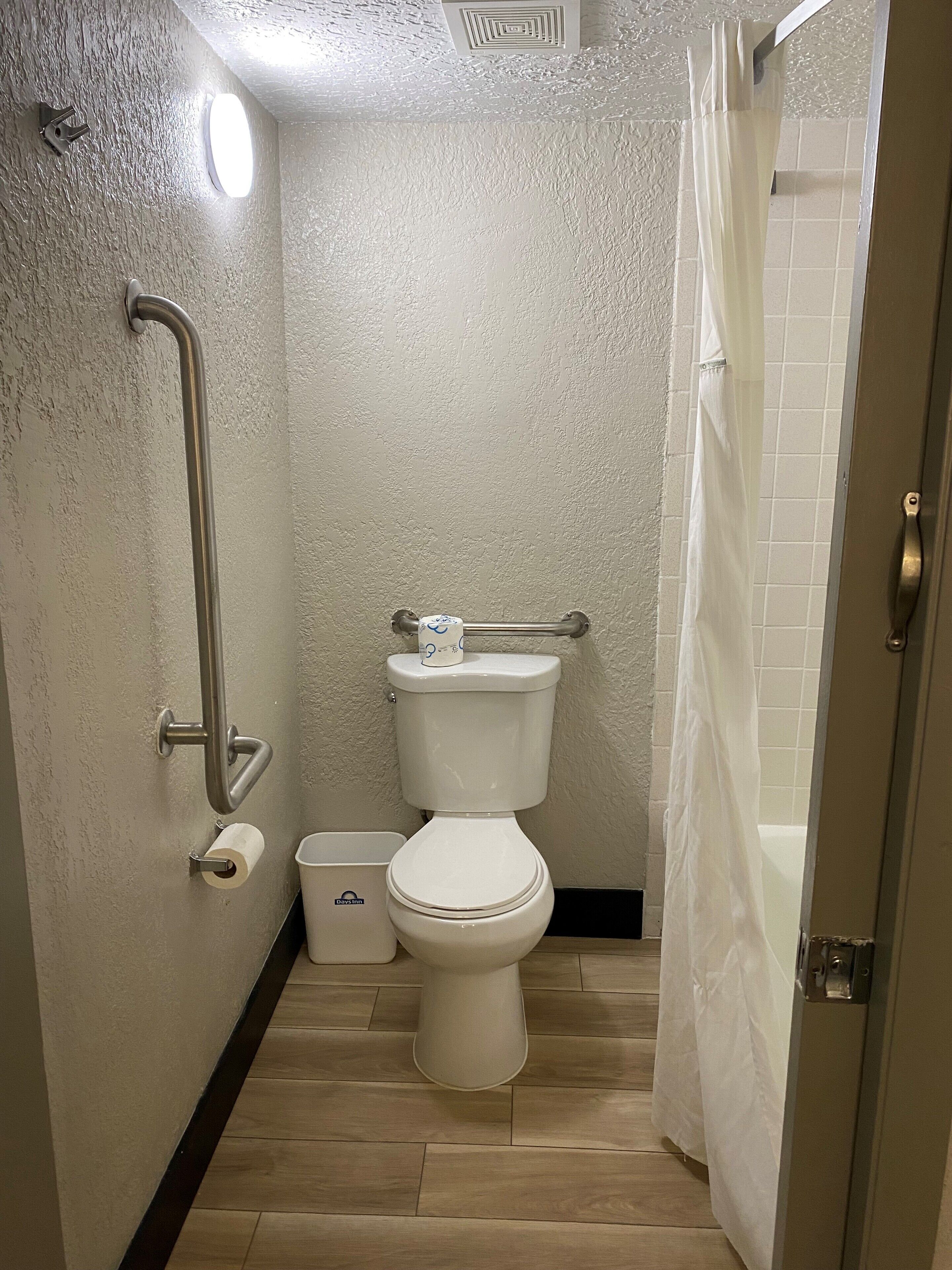 A small, compact bathroom with a white toilet, a handrail, a shower curtain, and a trash bin.