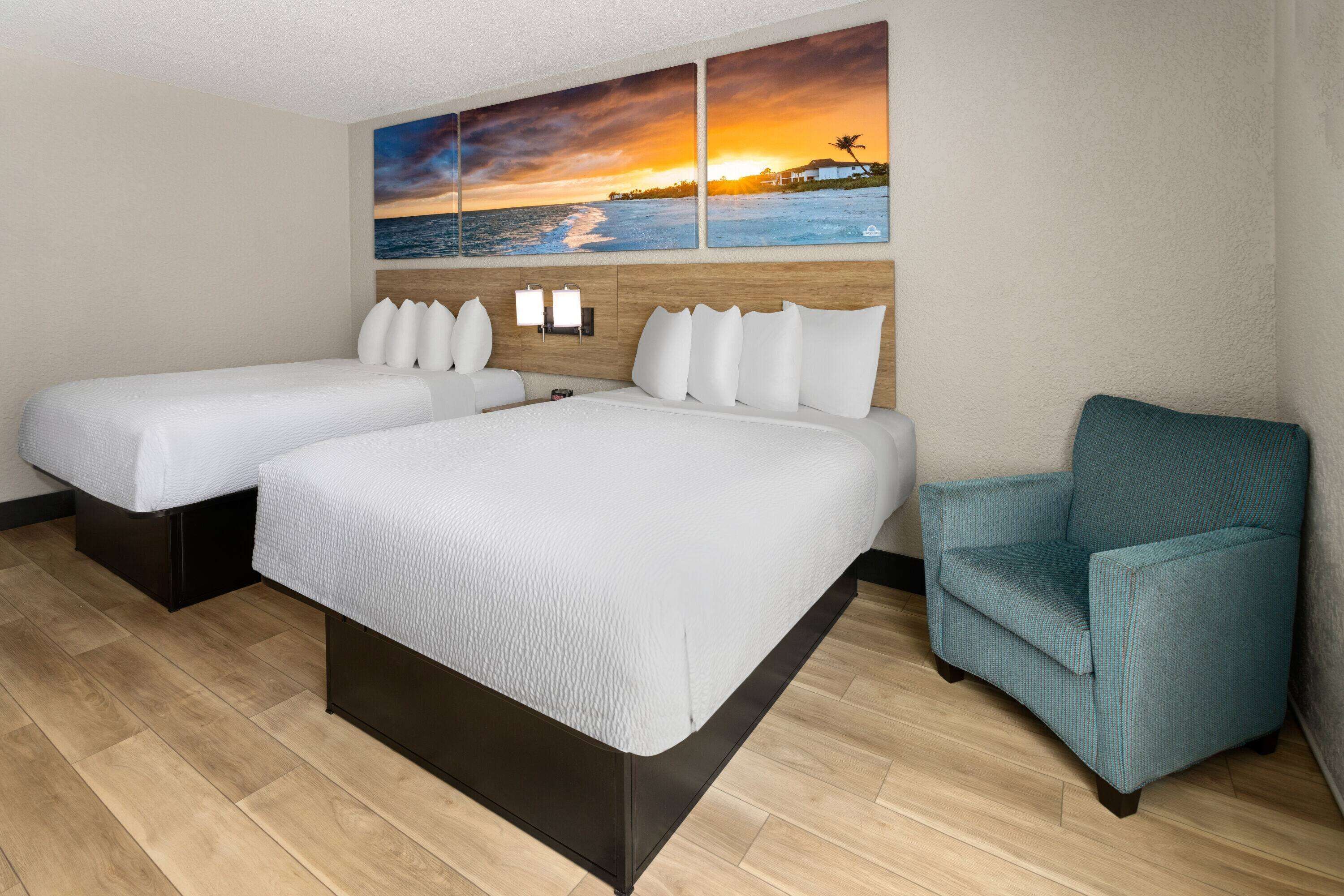 Une chambre d’hôtel avec deux lits, un fauteuil turquoise et un tableau représentant un coucher de soleil accroché au mur.
