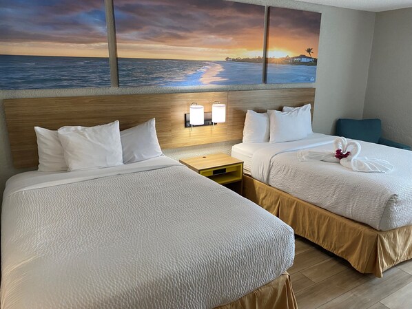 Habitación de hotel con dos camas, una mesita de noche, una lámpara de pared y una pintura con vista al atardecer.