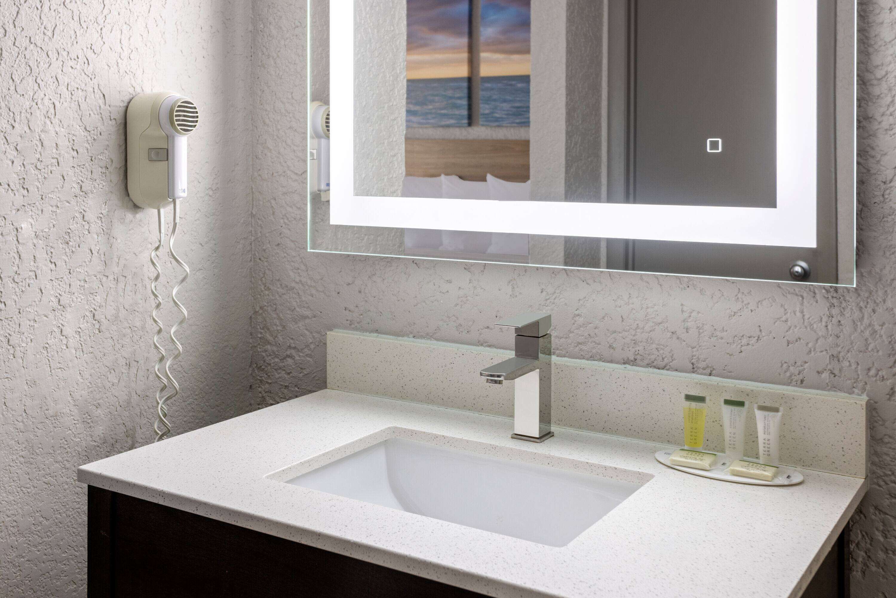 A bathroom with a white sink, a mirror with a view of the sea, and a hairdryer on the wall.