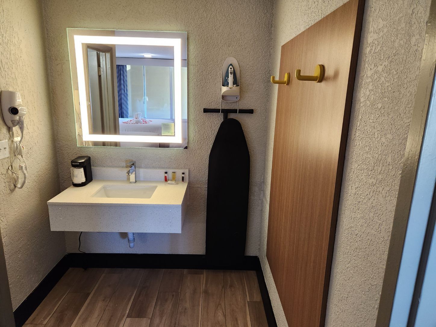 A bathroom with a white sink, a mirror with a light, a hairdryer, a coffee maker, and a wooden door.