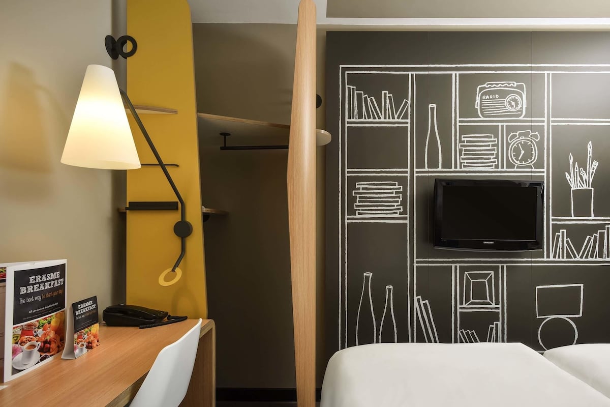 A hotel room with a desk, chair, lamp, bed, TV, and a chalkboard wall with drawings.