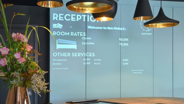 A reception desk with a digital display listing room rates and other services. - ibis Brussels Erasmus (Brussels)
