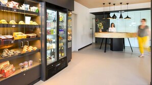A modern hotel lobby with a refrigerated display case filled with snacks and beverages, a reception desk, and pendant lighting. - ibis Brussels Erasmus (Brussels)