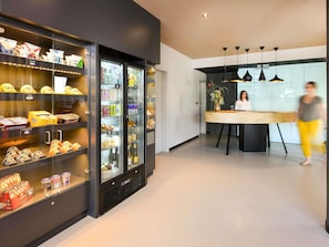 A modern hotel lobby with a refrigerated display case filled with snacks and beverages, a reception desk, and pendant lighting. - ibis Brussels Erasmus (Brussels)