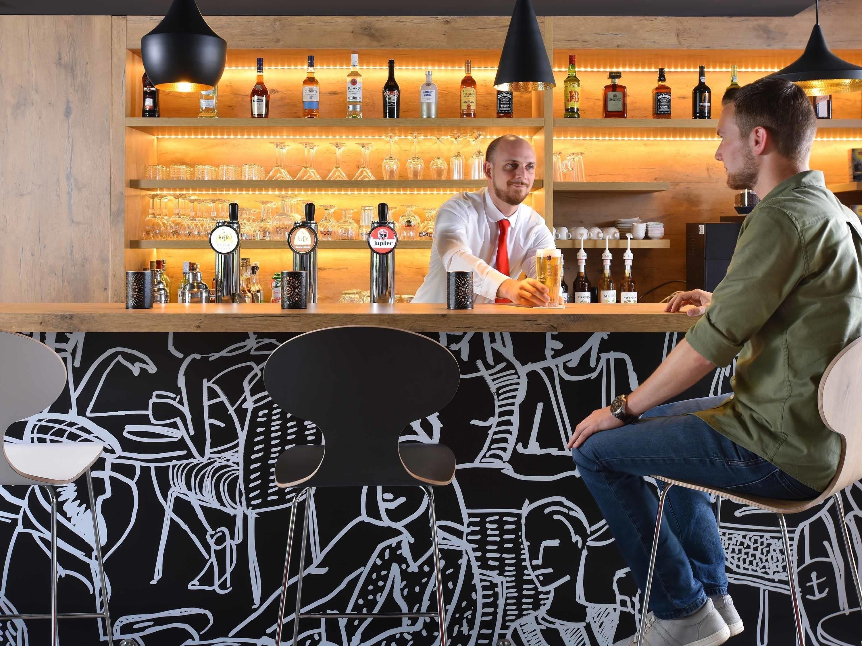 Een barruimte met een barman die een klant bedient. Achter de bar staan meerdere flessen en glazen op rekken.