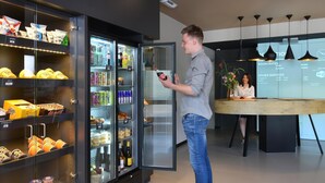 A man using a smartphone in a modern hotel lobby with a refrigerated display case filled with snacks and beverages. - ibis Brussels Erasmus (Brussels)