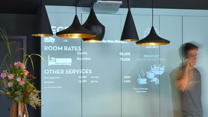 A hotel reception area with a wooden reception desk, black pendant lights, a vase with flowers, and a digital display listing room rates and services. - ibis Brussels Erasmus (Brussels)