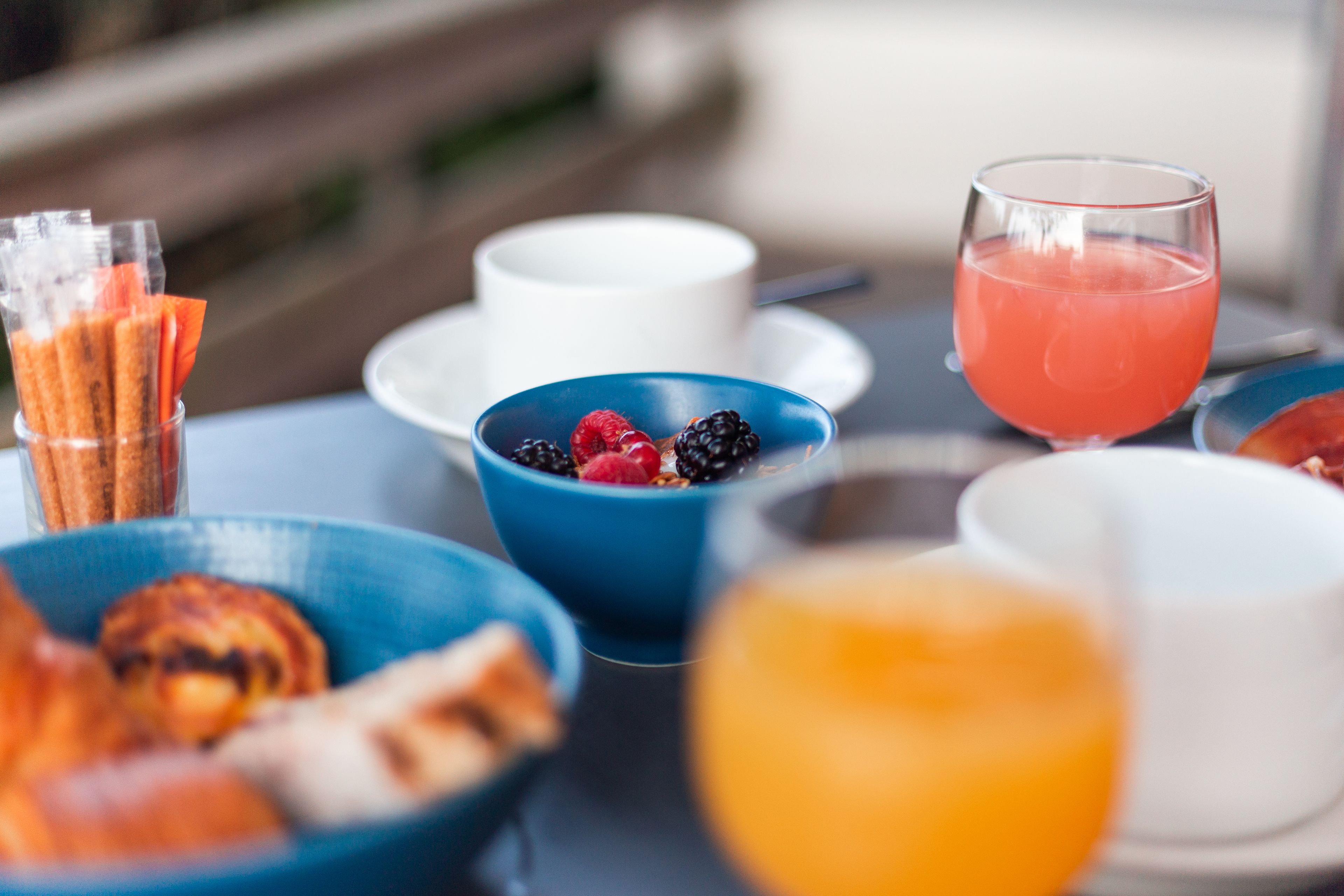 Una mesa de desayuno con varios platos, incluyendo un tazón azul con bayas, un vaso de jugo de naranja y una taza blanca.
