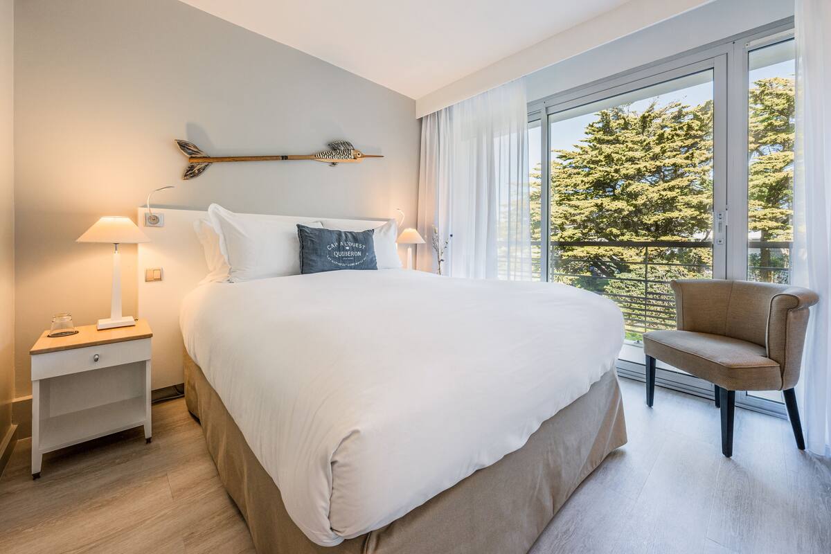 Ein Schlafzimmer mit einem großen Bett, Nachttischen, einem Sessel und einem Fenster mit Blick auf Bäume.