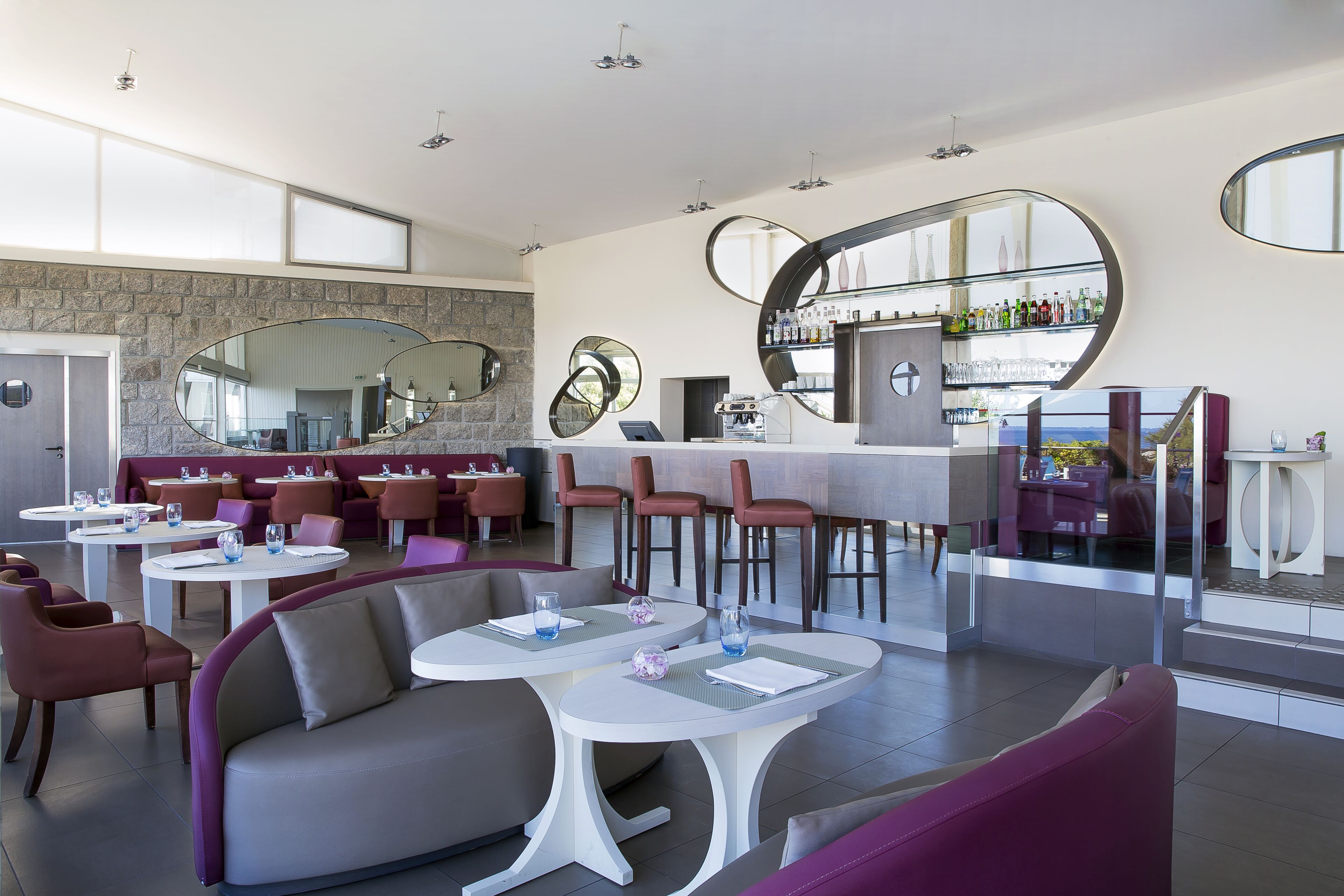 A modern restaurant with round tables, cushioned seating, and a bar area with a glass enclosure.
