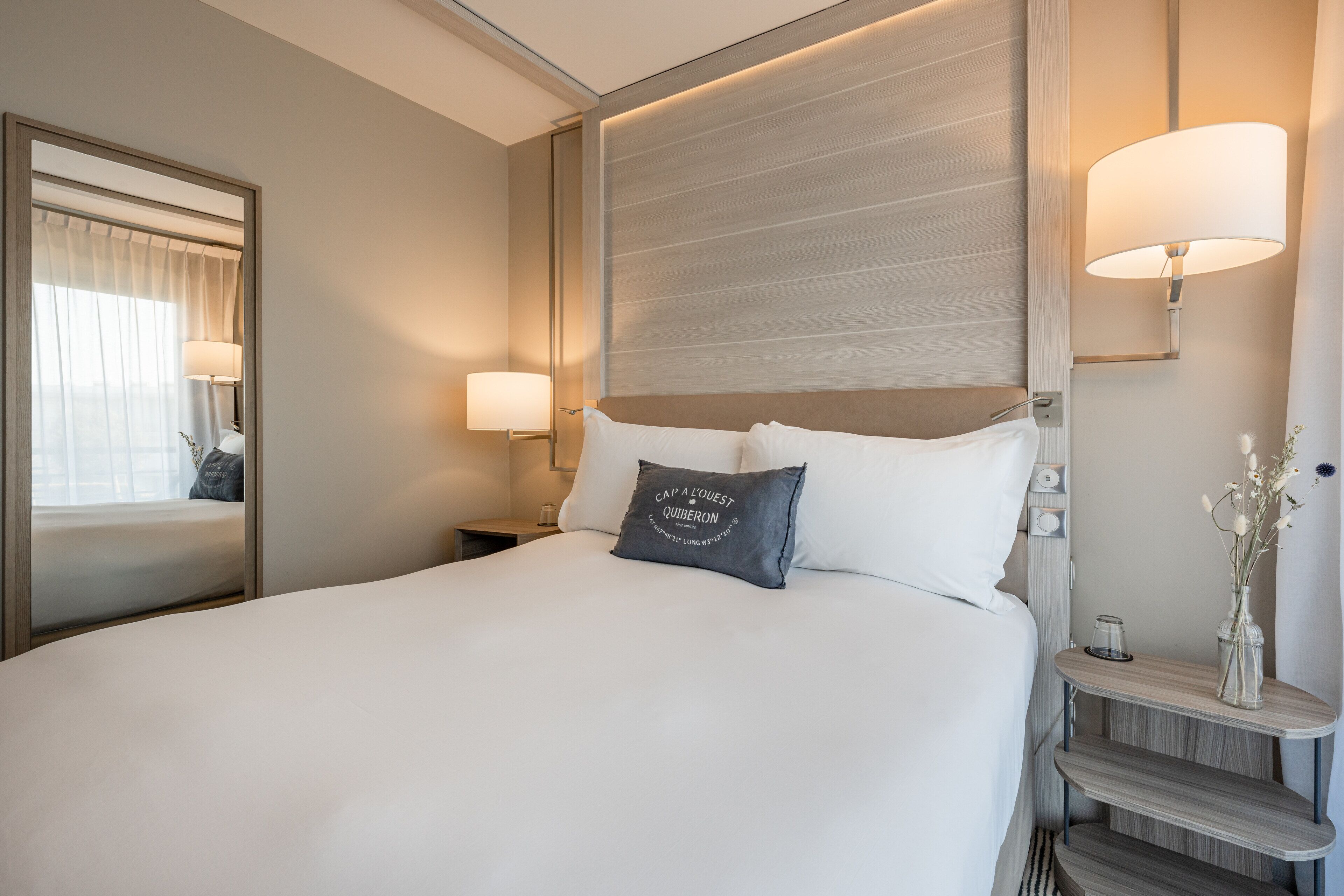 A neatly made bed with white linens, a dark gray pillow with text, a bedside table with a vase of flowers, and a wall-mounted lamp.
