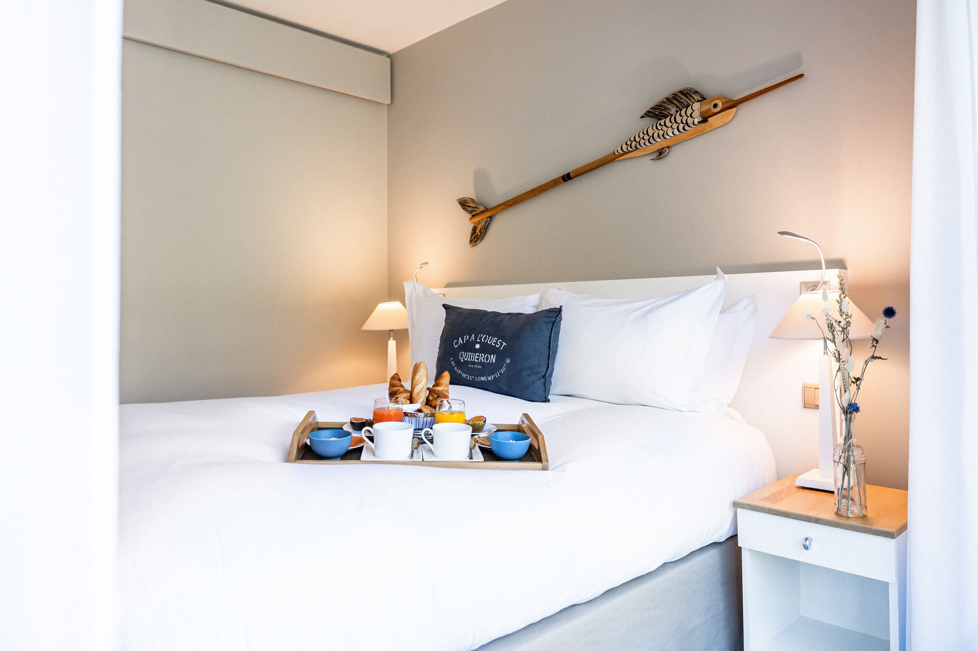 Una cama bien hecha con una bandeja de desayuno, una mesita de noche con lámpara y un objeto decorativo en la pared.