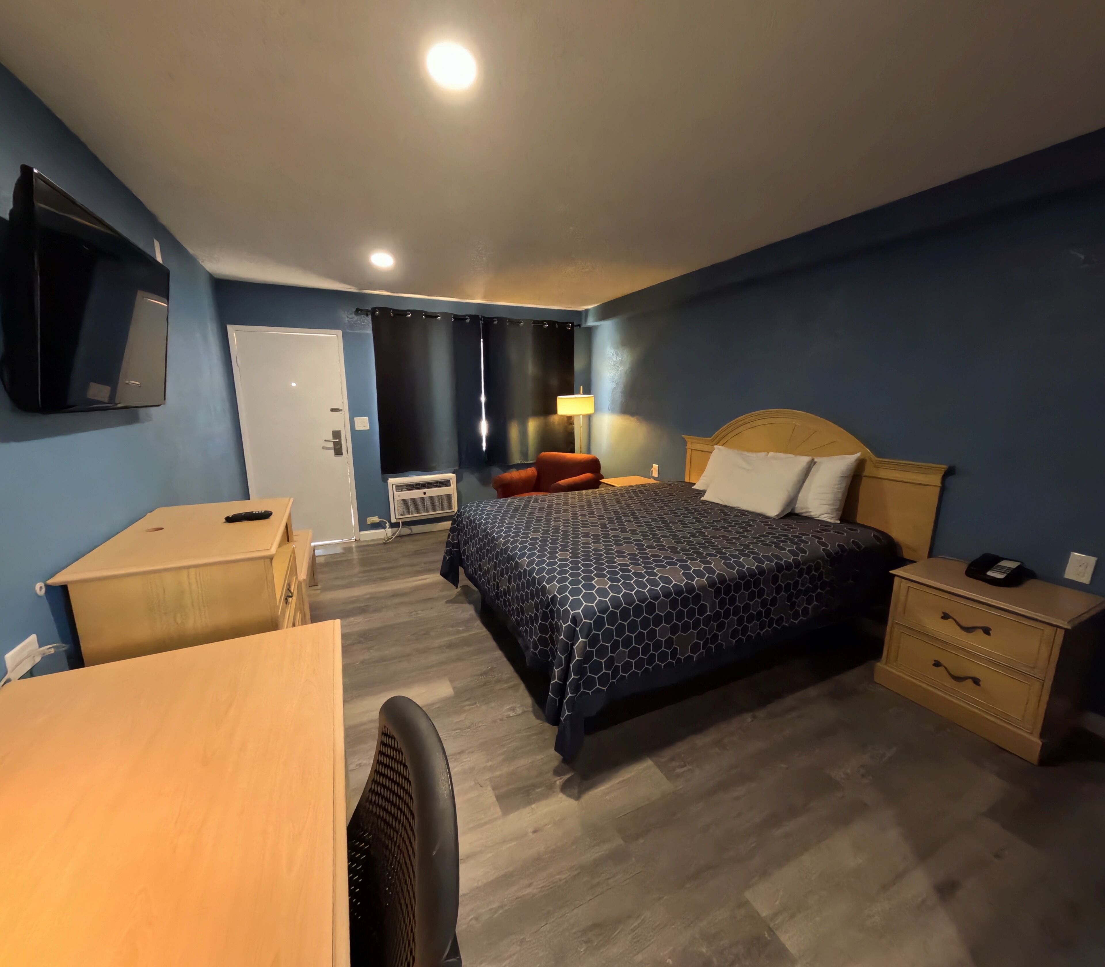 A hotel room with a bed, bedside table, desk, chair, TV, air conditioning unit, and a lamp.