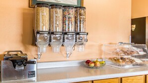Um balcão de pequeno-almoço com distribuidores de cereais, uma máquina de café e uma taça de fruta.