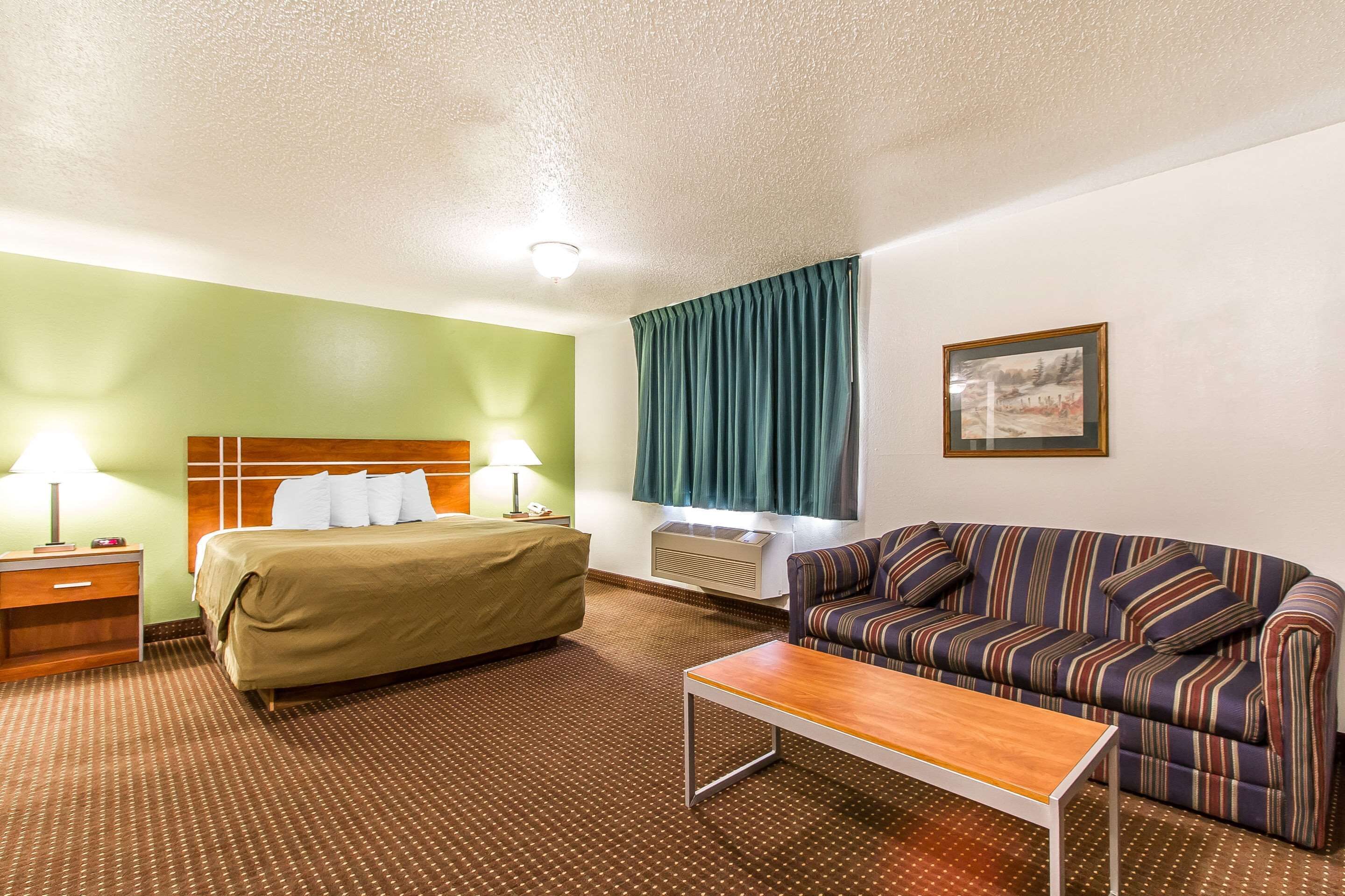 A hotel room with a bed, a sofa, a table, and a wall-mounted air conditioner.
