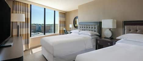 Habitación de hotel con dos camas, un televisor, un escritorio y un ventanal grande con cortinas.