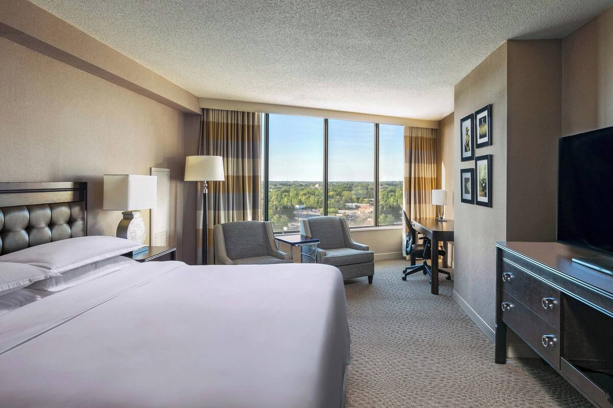 A hotel room with a large bed, a desk, two chairs, a dresser, and a TV.