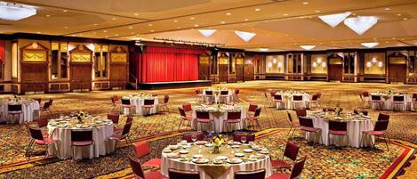 Un gran salón de banquetes con mesas y sillas redondas, un escenario rojo y candelabros ornamentados.