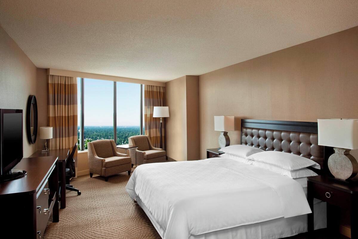 A hotel room with a large bed, two armchairs, a desk with a TV, and a view of the cityscape through large windows.