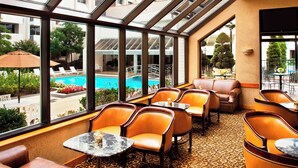Un área de descanso junto a la piscina con sillones de cuero y mesas, amplios ventanales con vistas a una piscina y vegetación.