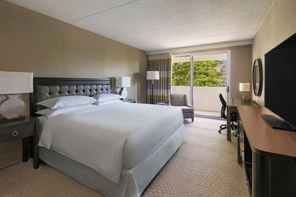 A hotel room with a large bed, two bedside tables with lamps, a desk with a chair, a TV, and a balcony with a view of trees.
