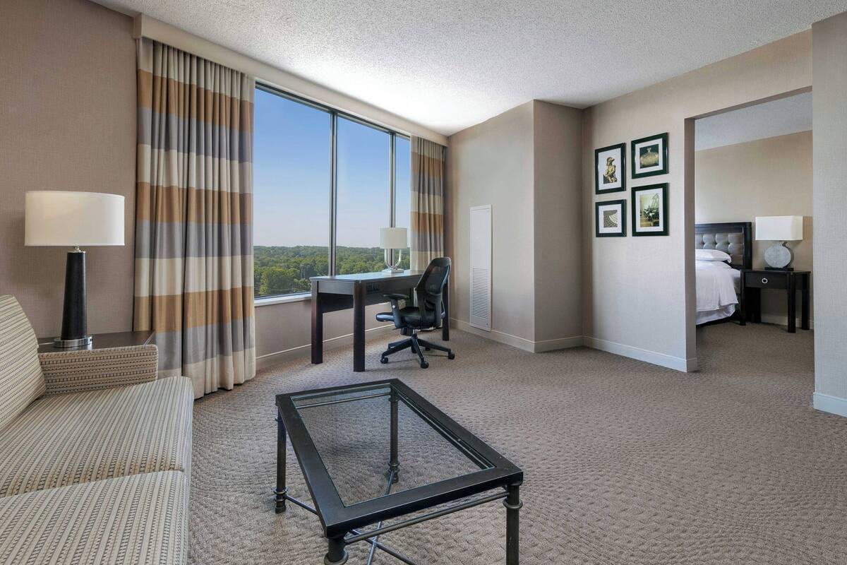 A hotel room with a large window, a desk, a chair, a bed, and a coffee table.