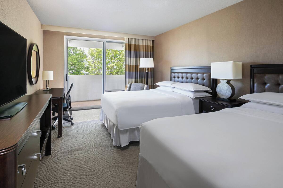 A hotel room with two beds, a desk, a chair, a TV, and a balcony with curtains.