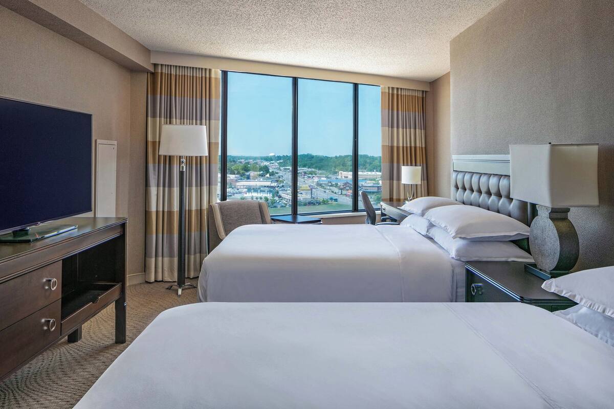 A hotel room with two beds, a TV, and a large window offering a city view.