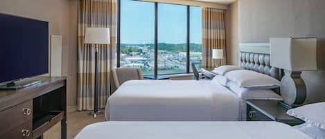 A hotel room with two beds, a TV, and a large window offering a city view.