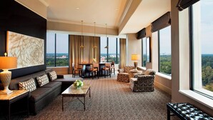 Amplia habitación de hotel con un ventanal que ofrece vistas a la ciudad, un sofá de color oscuro, una zona de comedor con mesa y sillas, y un sillón estampado.