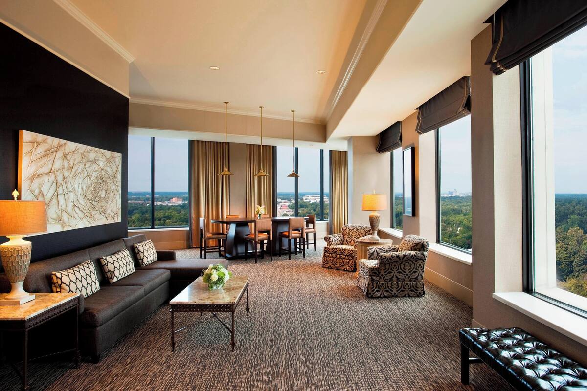A spacious hotel room with a large window offering a city view, a dark-colored sofa, a dining area with a table and chairs, and a patterned armchair.