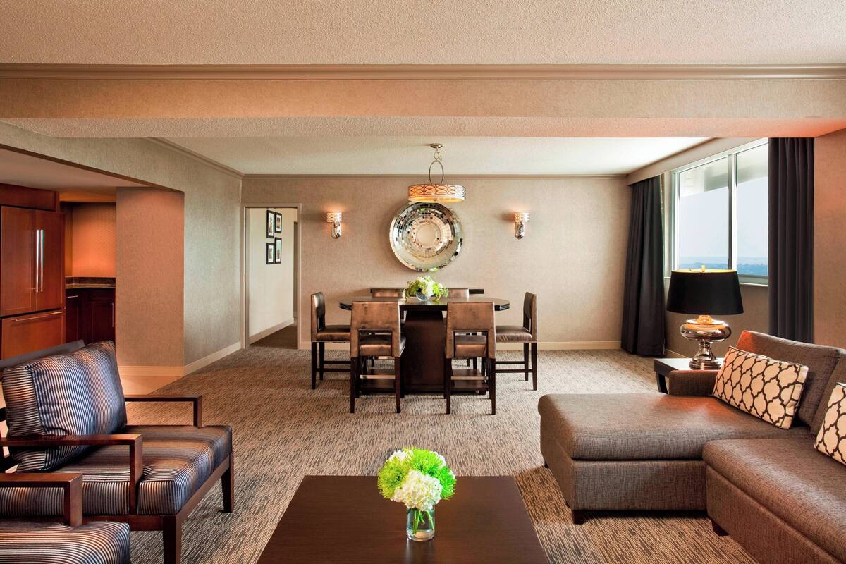 A hotel room with a dining area, a sofa, a coffee table with a vase, and a view of the city.