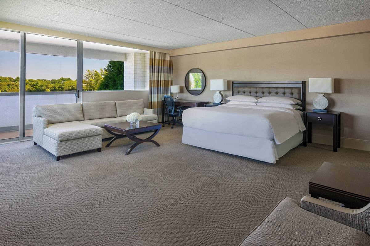 A hotel room with a large bed, a sofa, a desk, and a view of greenery through the window.