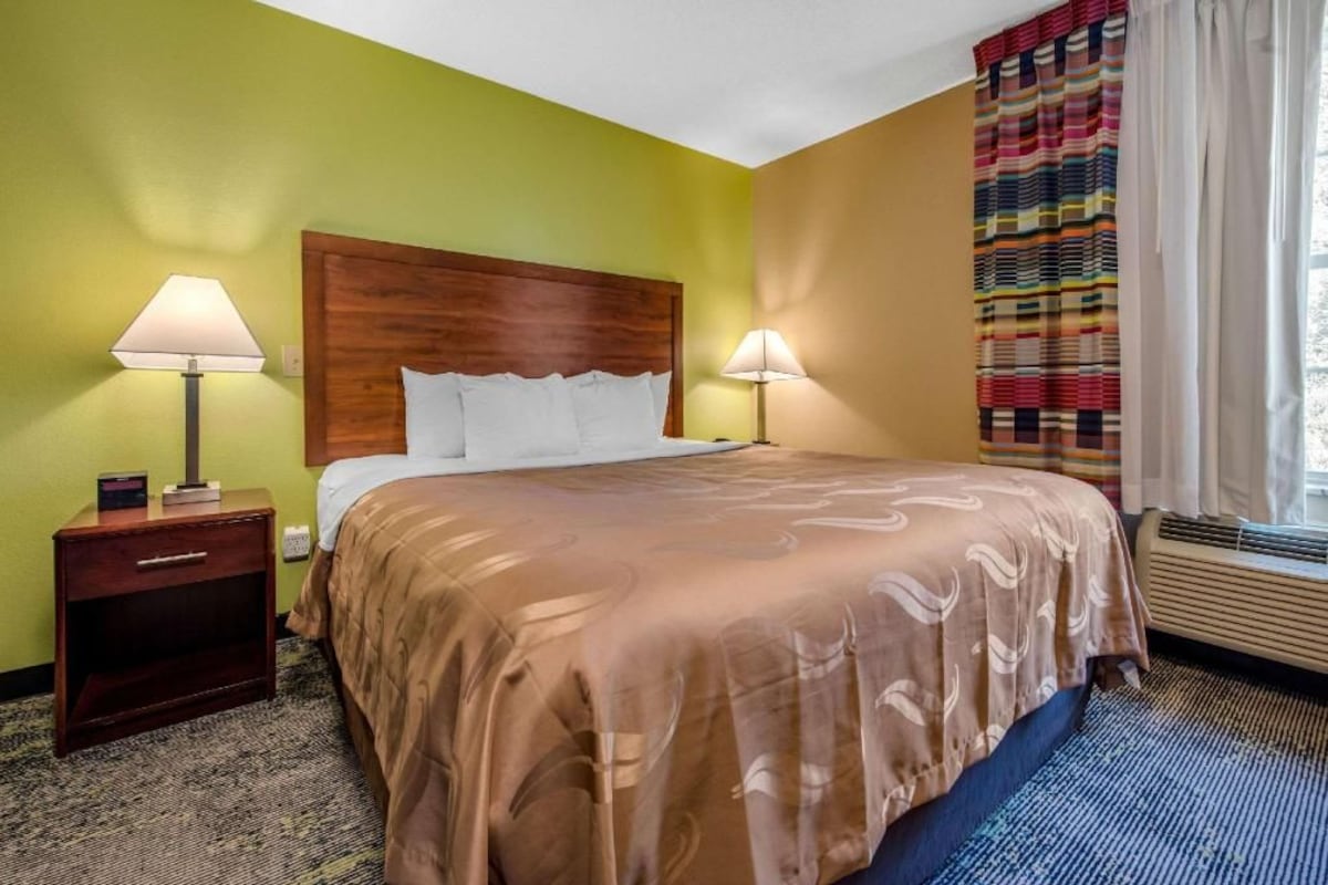 Une chambre d’hôtel avec un grand lit, deux tables de chevet avec des lampes, une fenêtre avec des rideaux et un sol recouvert de moquette.