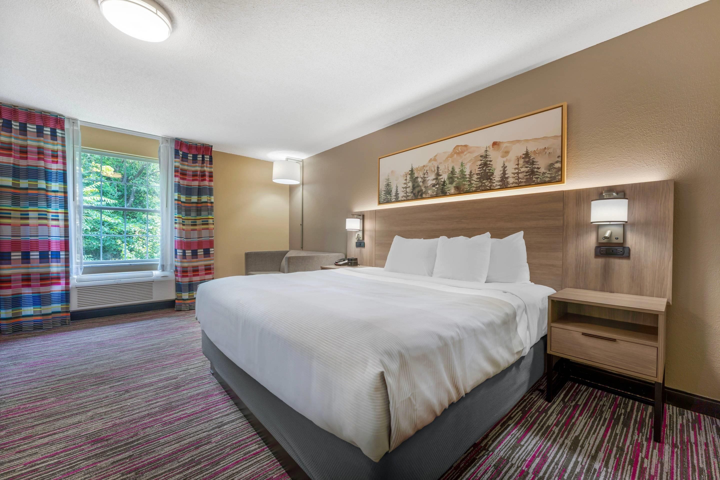 A hotel room with a large bed, a desk, a chair, a window with curtains, a painting on the wall, and a carpeted floor.