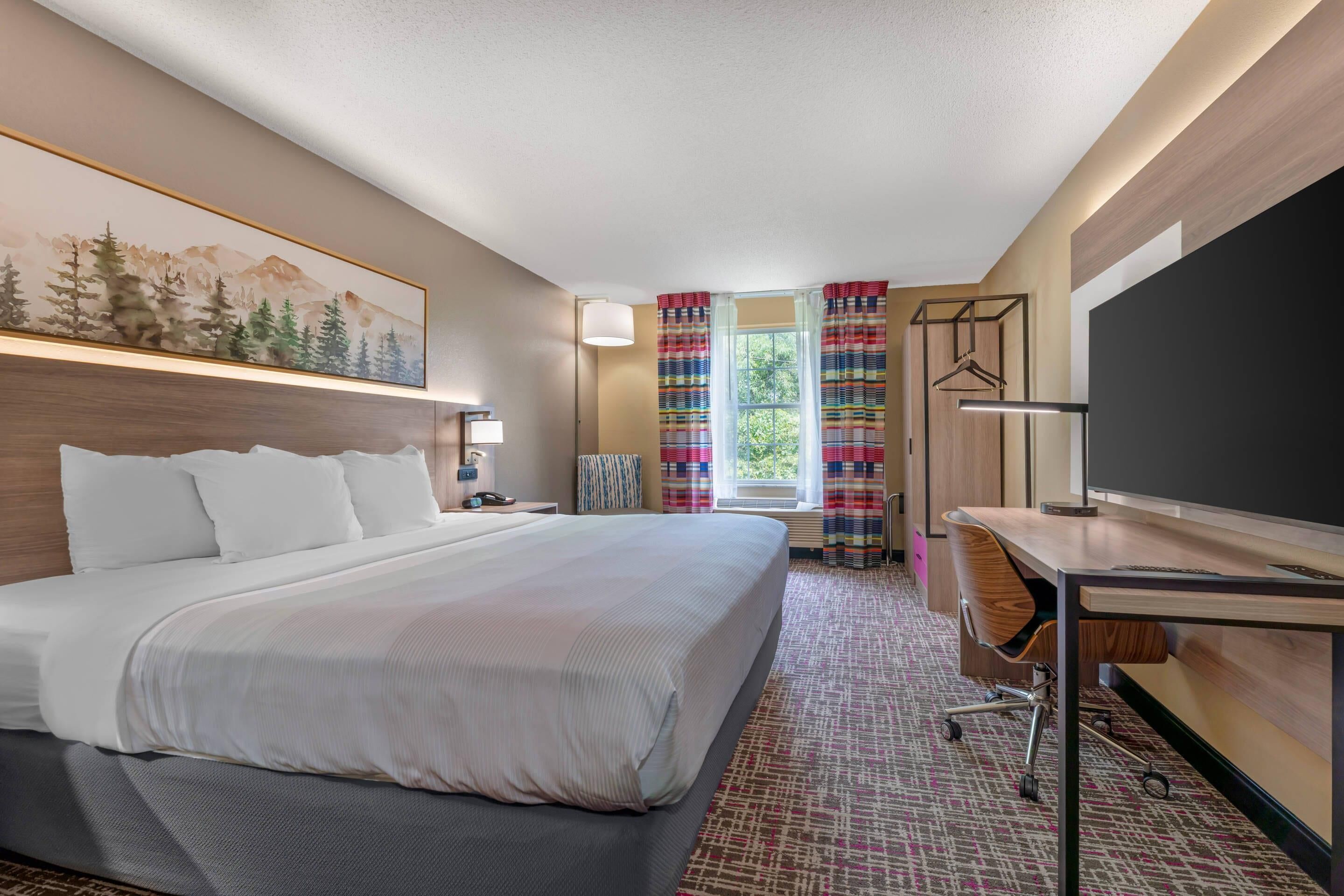 A hotel room with a large bed, a desk with a flat-screen TV, a coat rack, and a window with curtains.